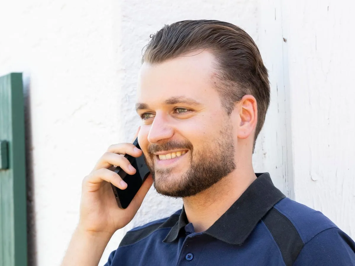 Felix Bernhauser telefoniert lachend vor einer weißen Hauswand mit grünen Fensterläden.