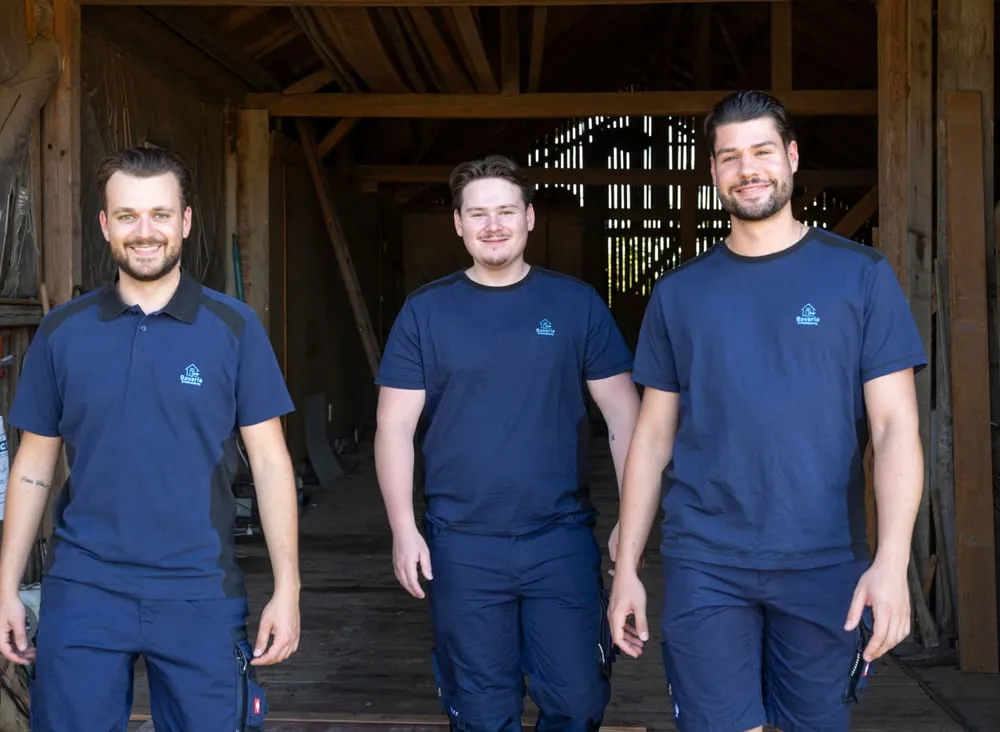 Felix Bernhauser, Mario Huber und ein Mitarbeiter in dunkelblauer Arbeitskleidung stehen lächelnd vor einer Scheune.